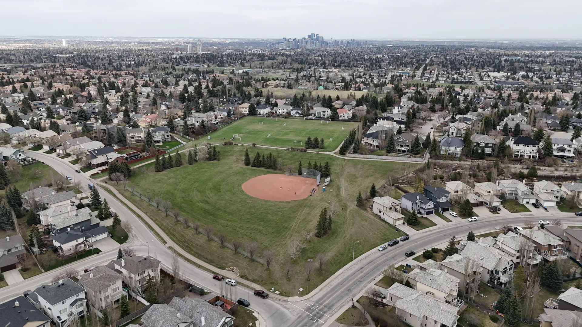 Aerial drone view of a Signal Hill community park with baseball diamond, soccer field, and residential streets with the Calgary downtown skyline on the horizon with Springbank Hill and Aspen Woods neighbourhoods visible in the distance