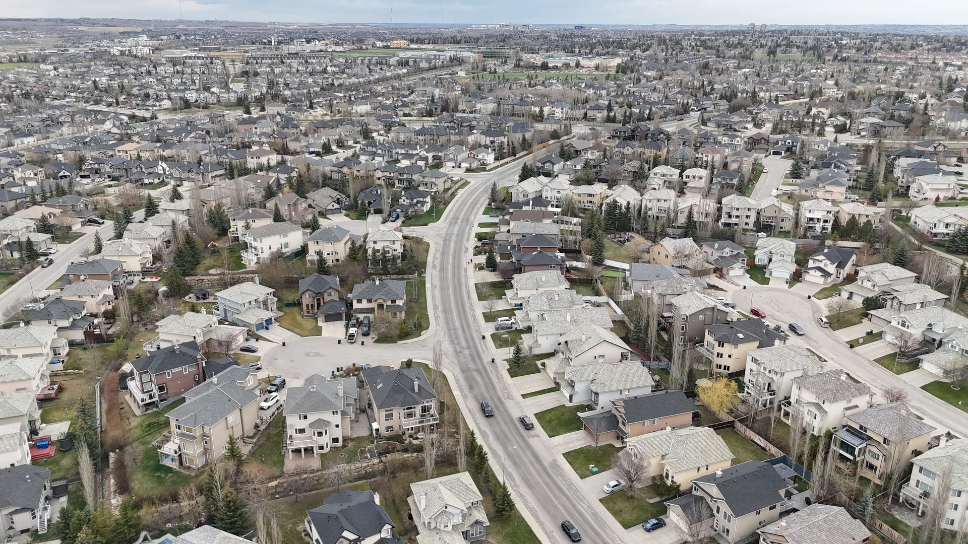 Aerial drone view of a main road through Signal Hill Calgary with established residential homes and mature neighbourhoods on both sides, looking east toward the city