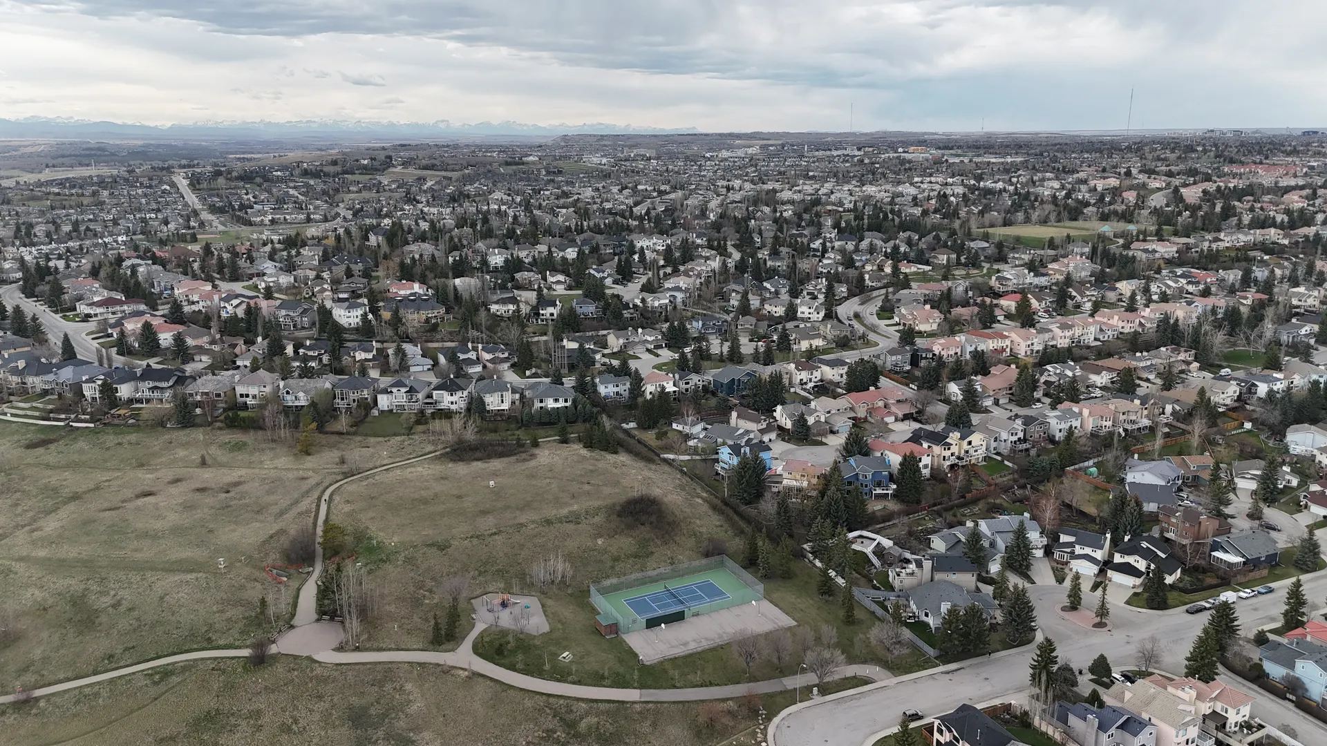 Aerial drone view of Signal Hill Calgary neighbourhood looking north with the Rocky Mountains on the horizon, residential streets, mature trees, and a community park with tennis courts in the foreground