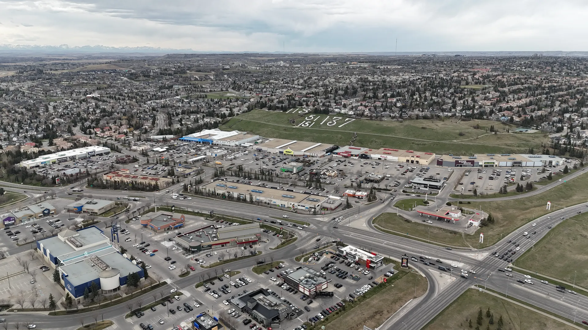 Aerial drone view of Westhills Towne Centre and Signal Hill commercial district with retail stores, parking lots, Battalion Park numerals, and the Rocky Mountains on the horizon